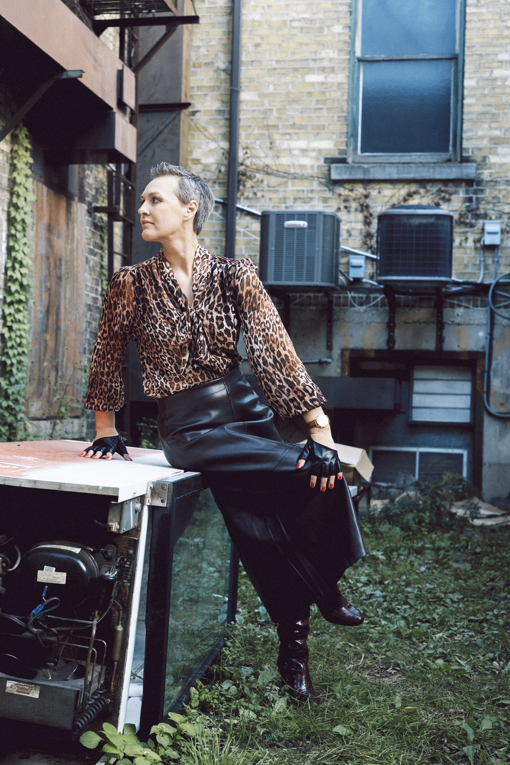 A stylish adult woman with short grey hair sits on a rusty appliance outdoors, wearing a leopard print top, black leather skirt, and boots.