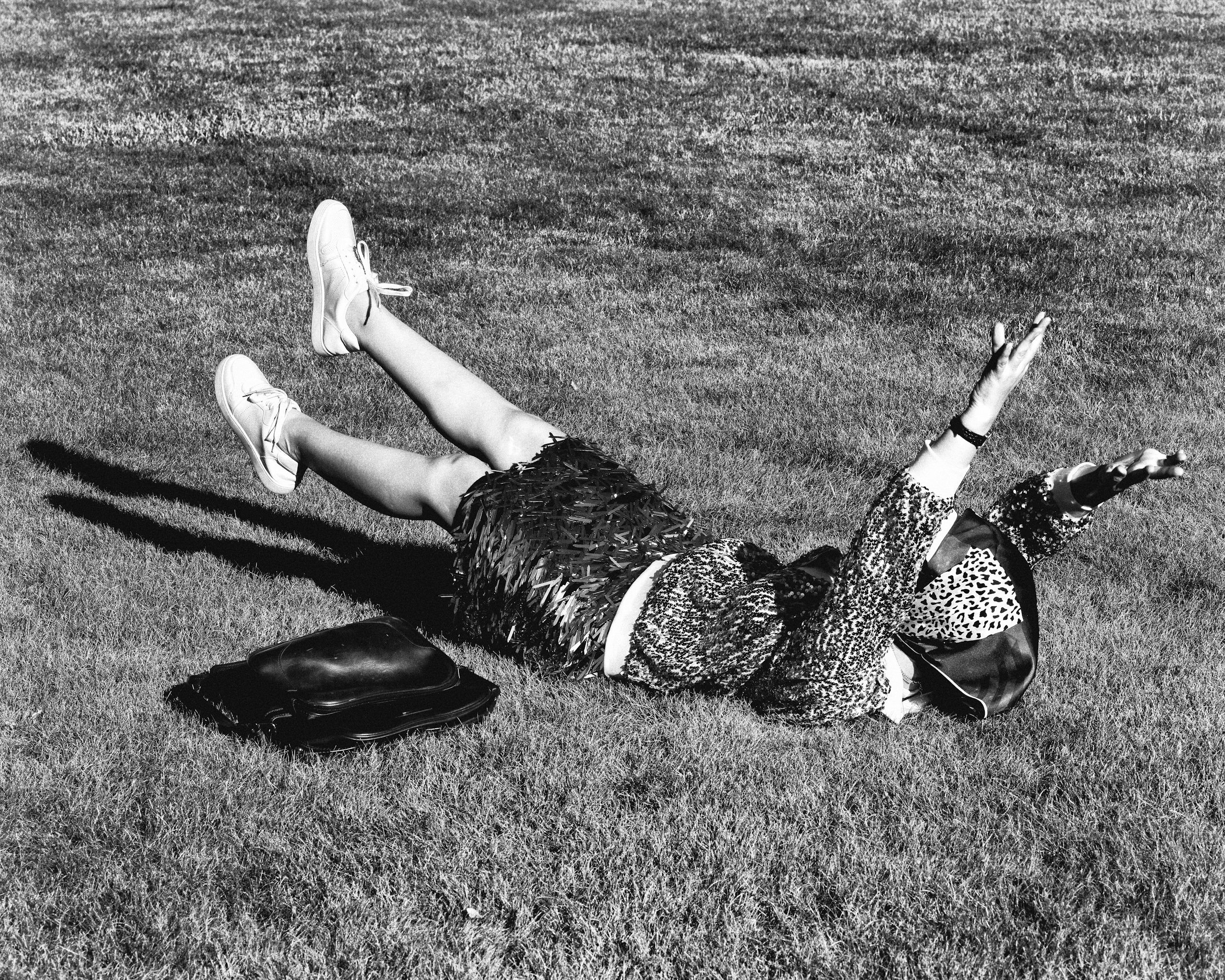 Woman in patterned dress and sneakers reclines on grass with arms raised, purse nearby in a black and white outdoor scene.