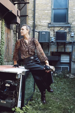Woman with short hair in a leopard print blouse and long black leather skirt sitting outdoors against a brick building.
