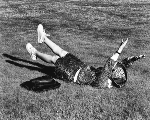 Monochrome shot of a woman in a textured skirt and white sneakers lying on a grass lawn with legs raised, a black bag beside her.