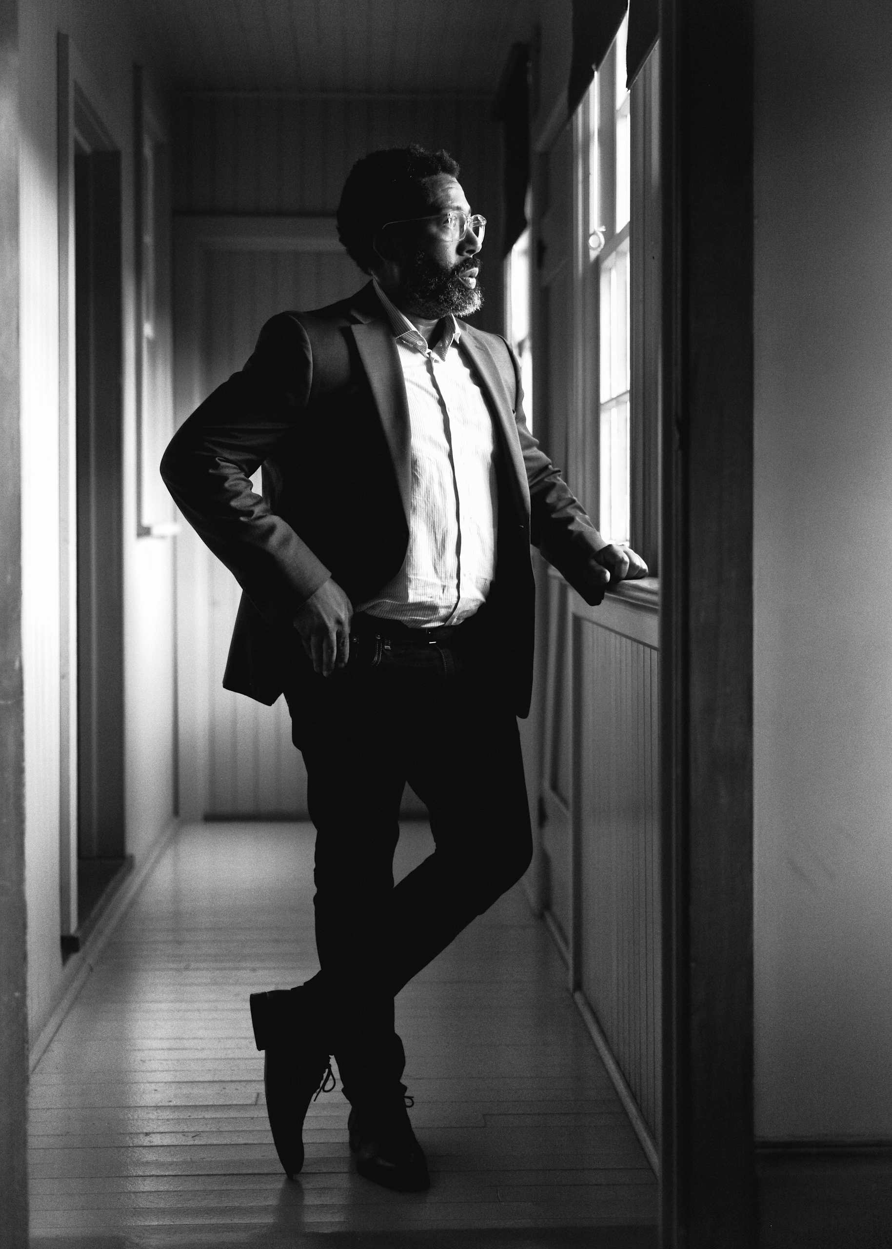 Black and white portrait of a man in a suit looking out a window in a hallway with natural light, creating a thoughtful and stylish image.