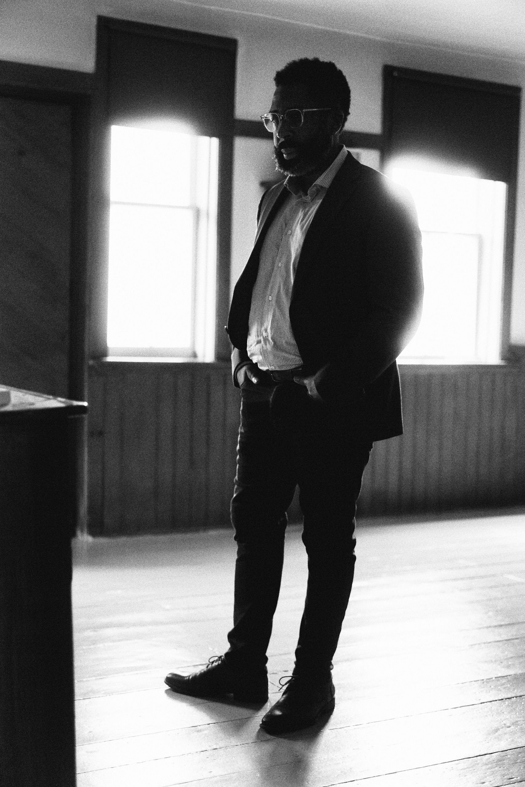 Full-length black-and-white studio shot of a stylish African-American man in a suit, posing against a bright, window-lit backdrop.