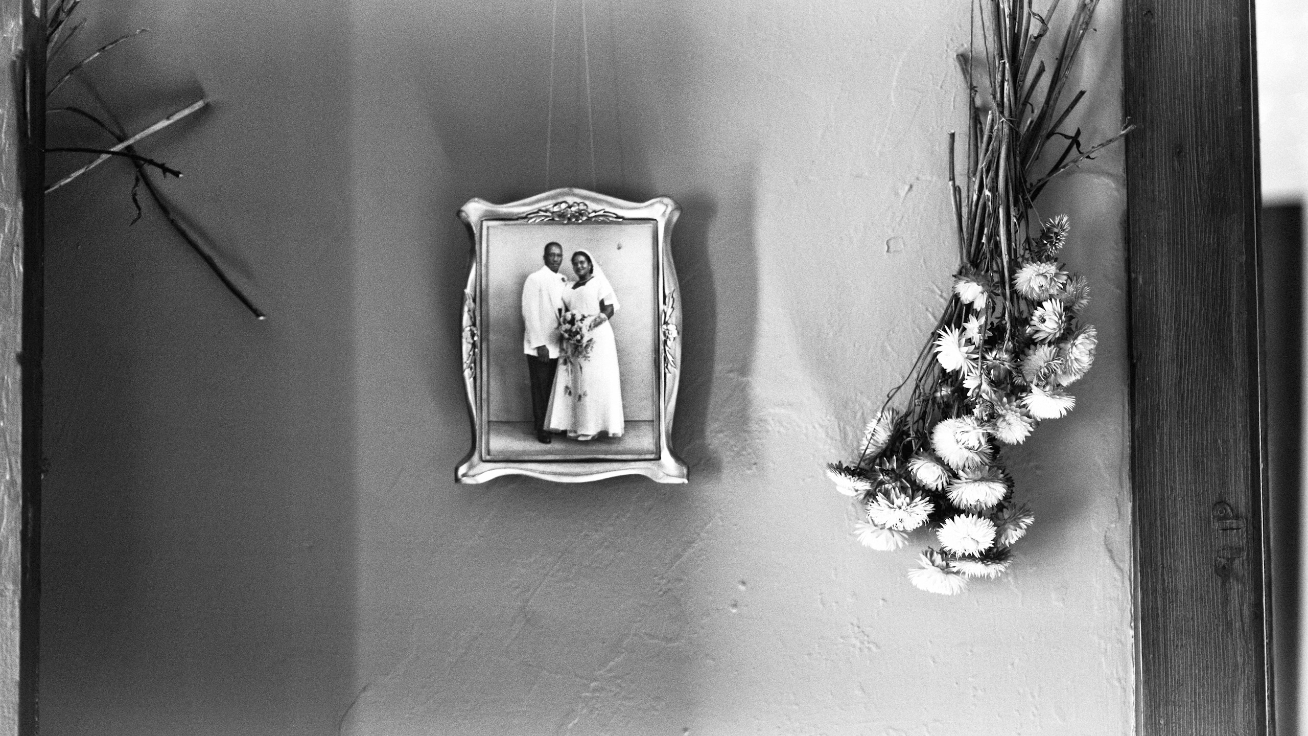 Monochrome photograph of a framed wedding picture hanging on a wall, complemented by dried flowers and branches for a rustic aesthetic.