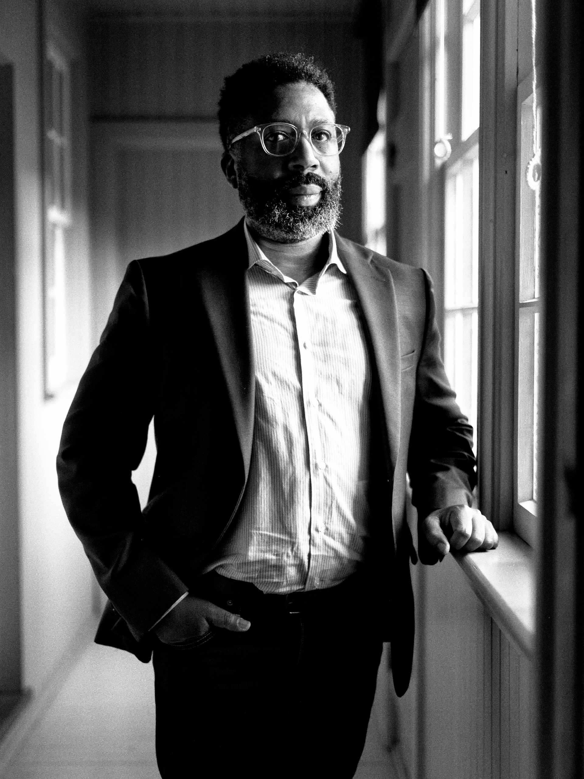 Black and white portrait of a Black man with glasses and a beard standing beside a window in a hallway wearing a dark blazer.
