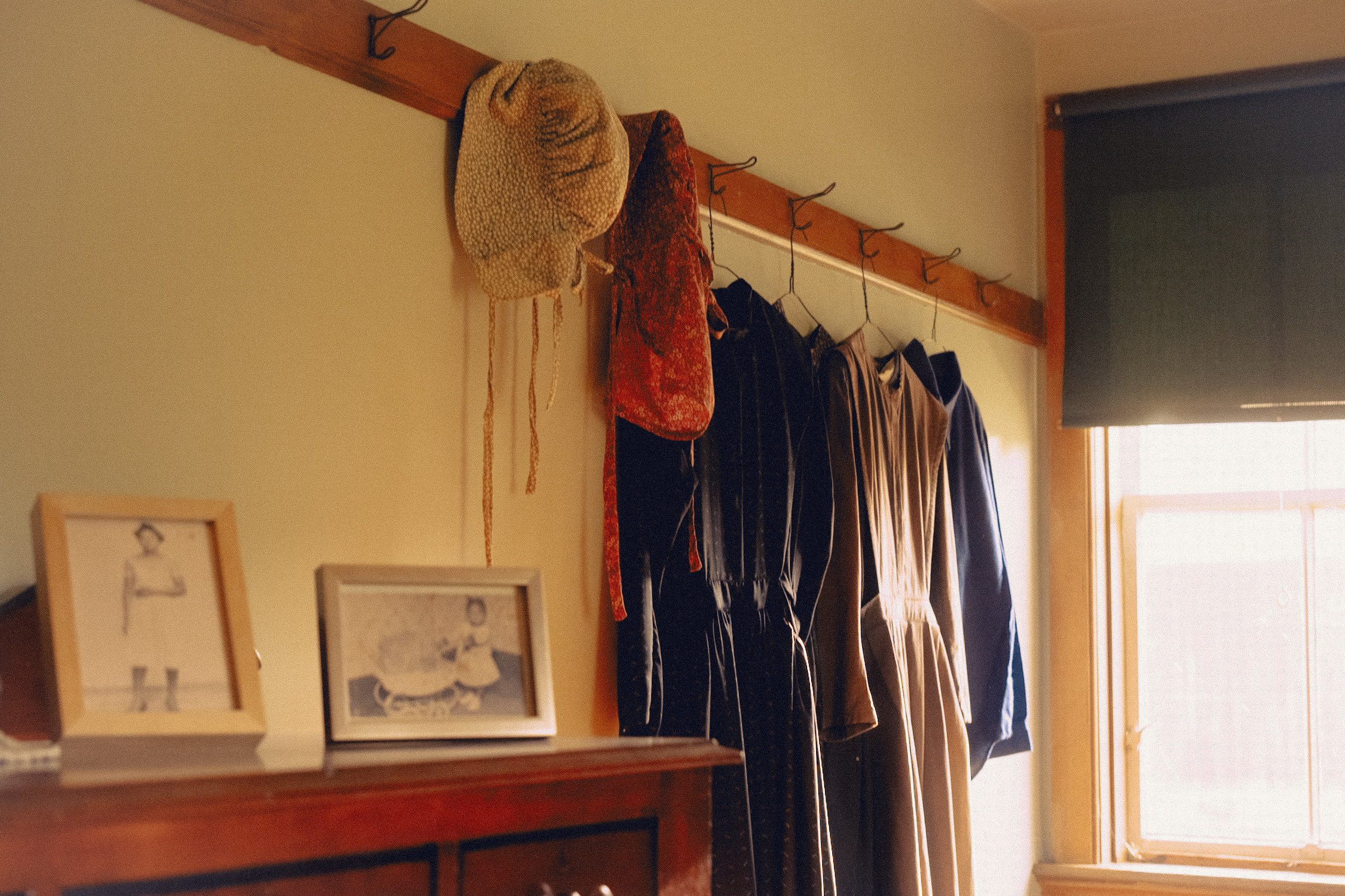 Vintage dresses hang on hooks against a pale wall next to framed photos on a wooden dresser, creating a nostalgic indoor scene.