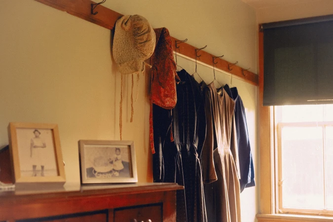 Vintage dresses hang on hooks against a pale wall next to framed photos on a wooden dresser, creating a nostalgic indoor scene.