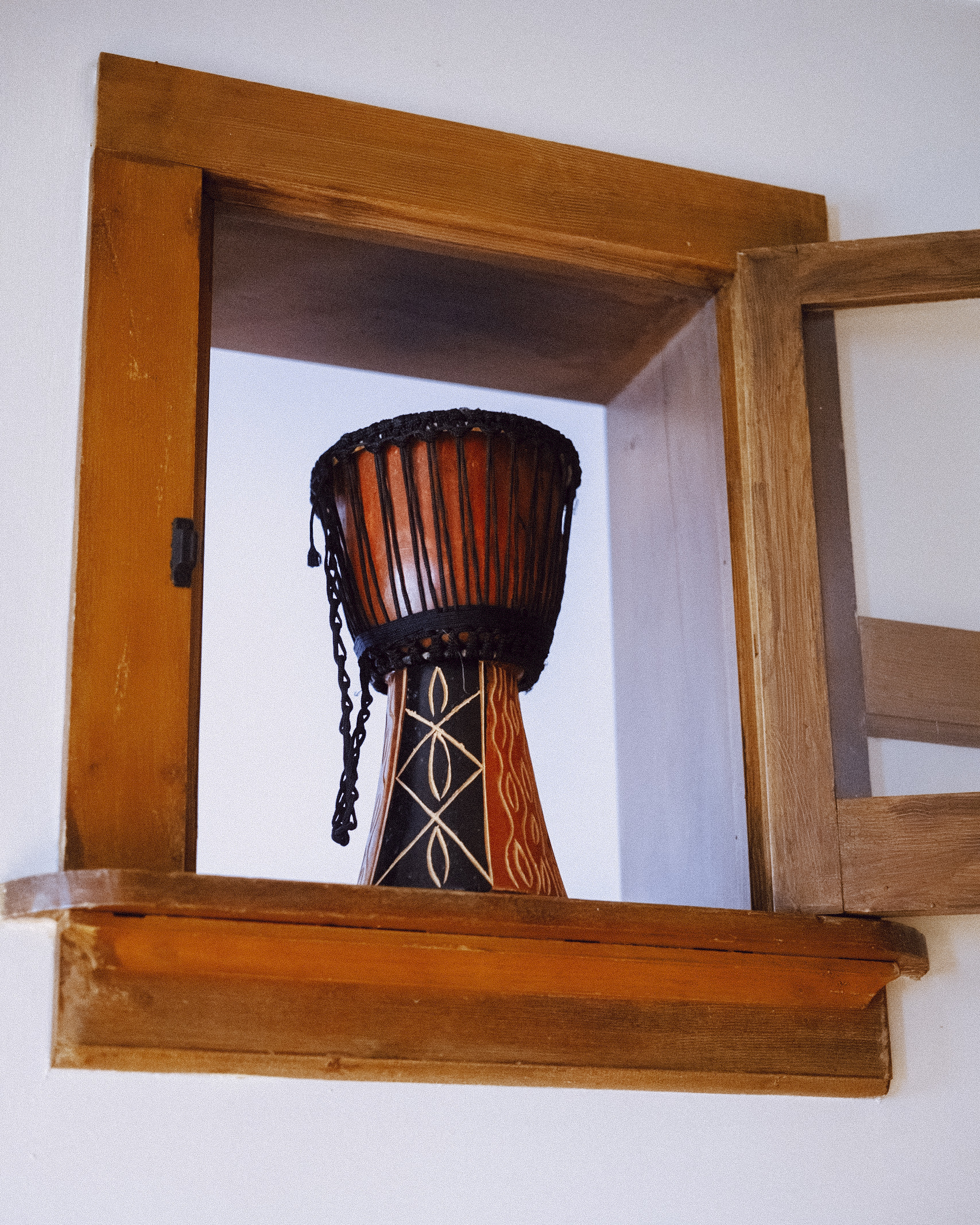 Close-up of a Djembe drum, decorated with geometric patterns, sitting inside of a rustic wooden window frame against a white wall.