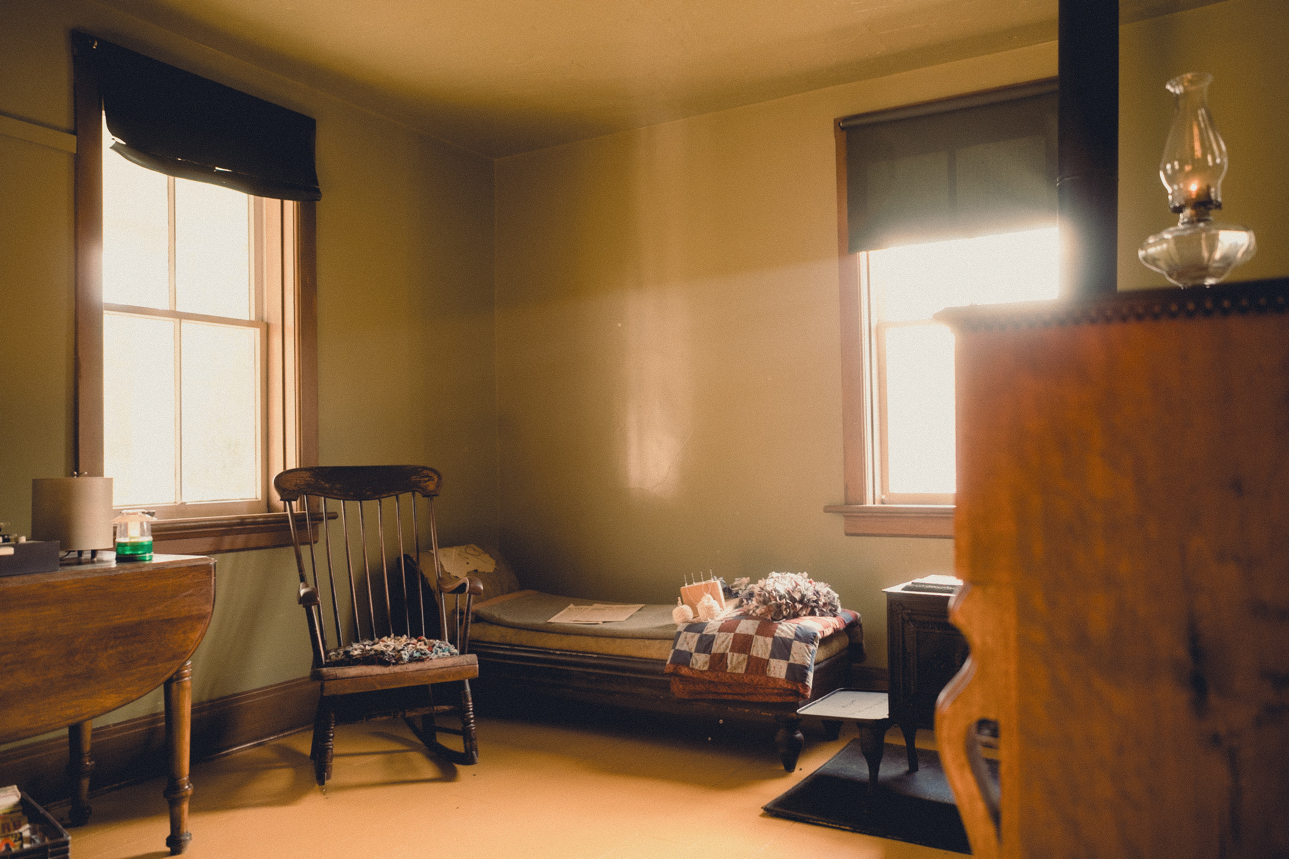 Antique bedroom featuring a wooden bed with a quilt, rocking chair, windows with dark shades, and a vintage lamp, creating a cozy, old-fashioned atmosphere.