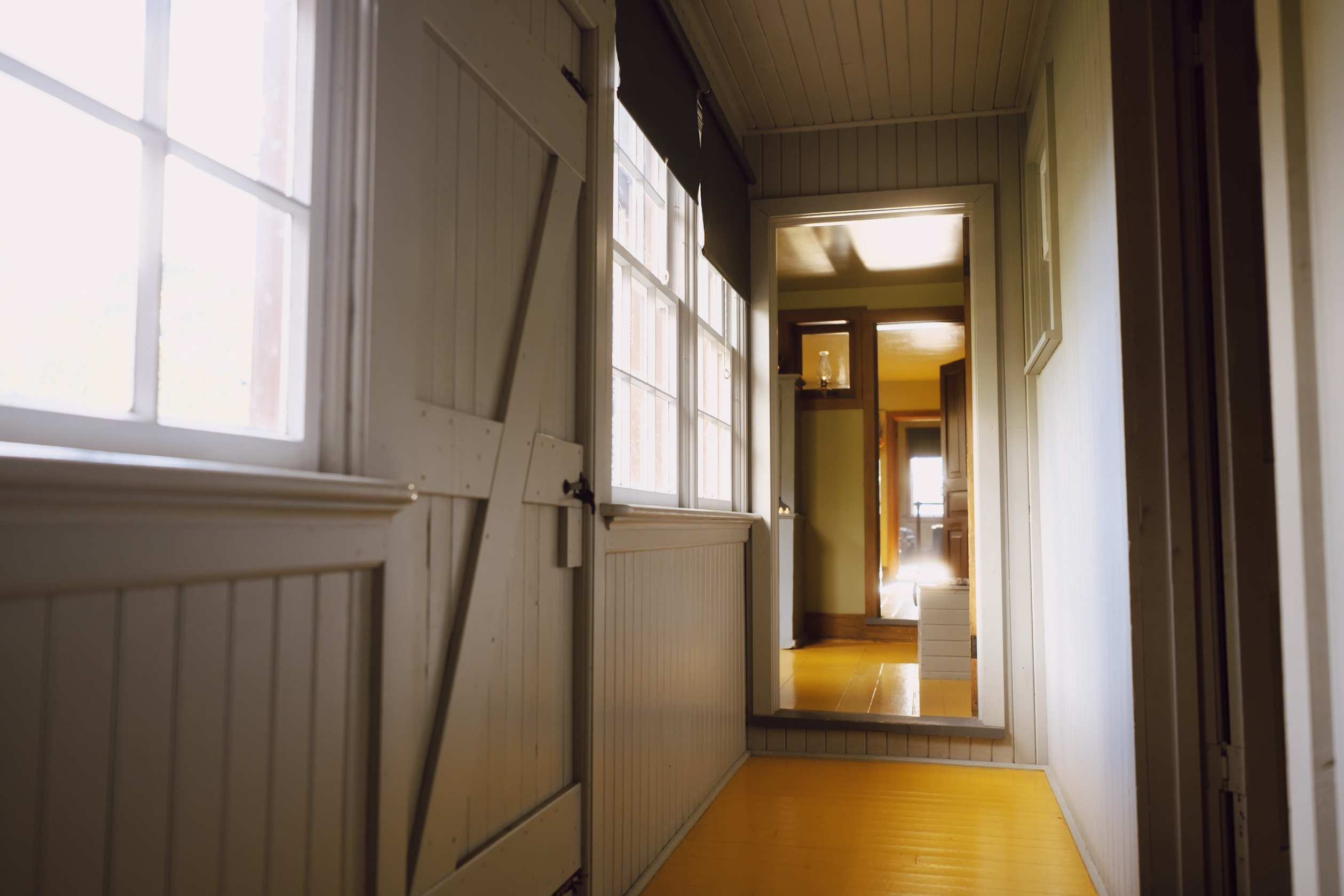 Indoor hallway with white wood paneling, doors, and windows, leading to rooms in the distance, flooded with bright natural light.