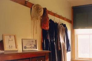 Historical dresses and cloth bonnets hanging from a wooden wall peg rail above framed vintage photographs on a wooden dresser.
