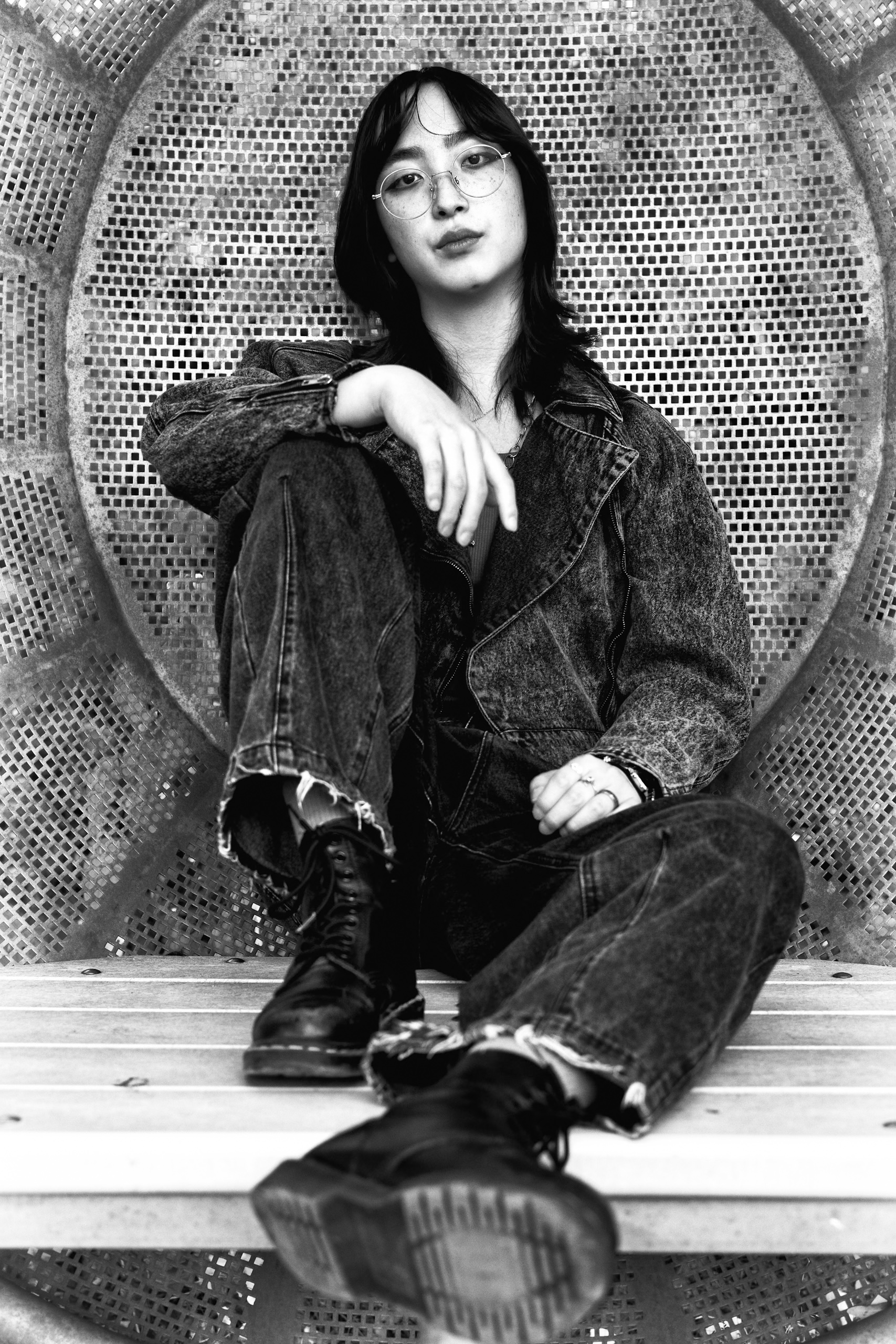 Young woman with glasses sits in denim jacket and boots in a gritty urban setting, leaning against a textured metallic wall.