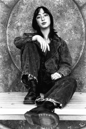 Young woman with glasses sits in denim jacket and boots in a gritty urban setting, leaning against a textured metallic wall.