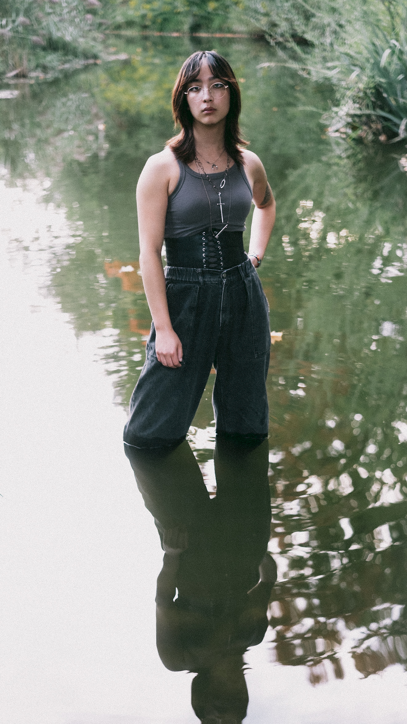 Young woman standing in water wearing a gray tank top and dark culottes with a cinched waist, reflective water, trees, and foliage.