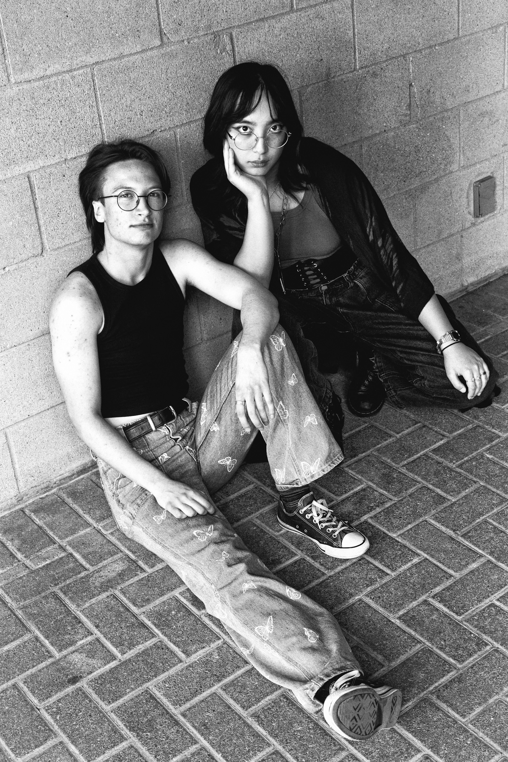 Black and white portrait of a non-binary couple sitting on a brick surface. The man wears jeans with butterflies and the woman a dress.