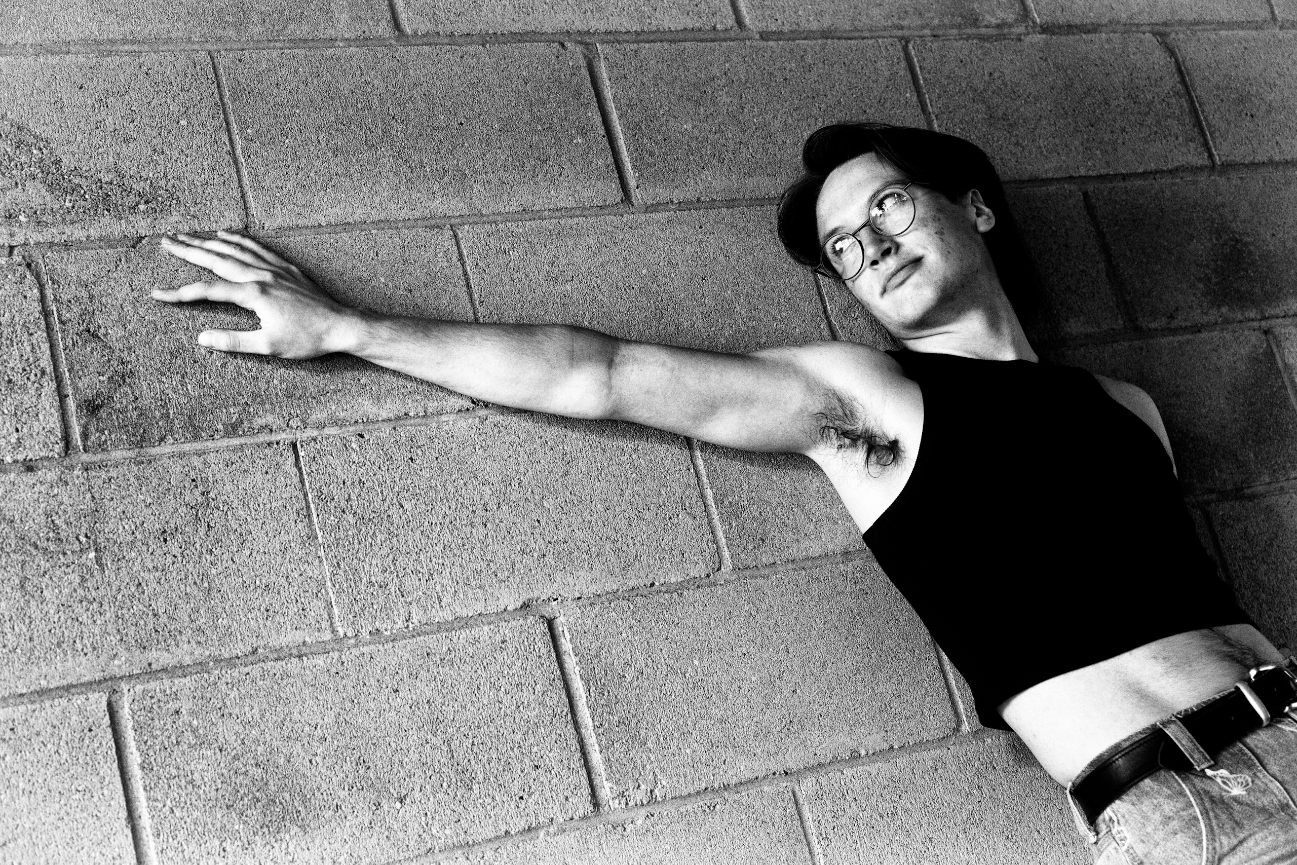 Black-and-white portrait of a man wearing glasses, a tank top, and jeans, leaning against a brick wall.