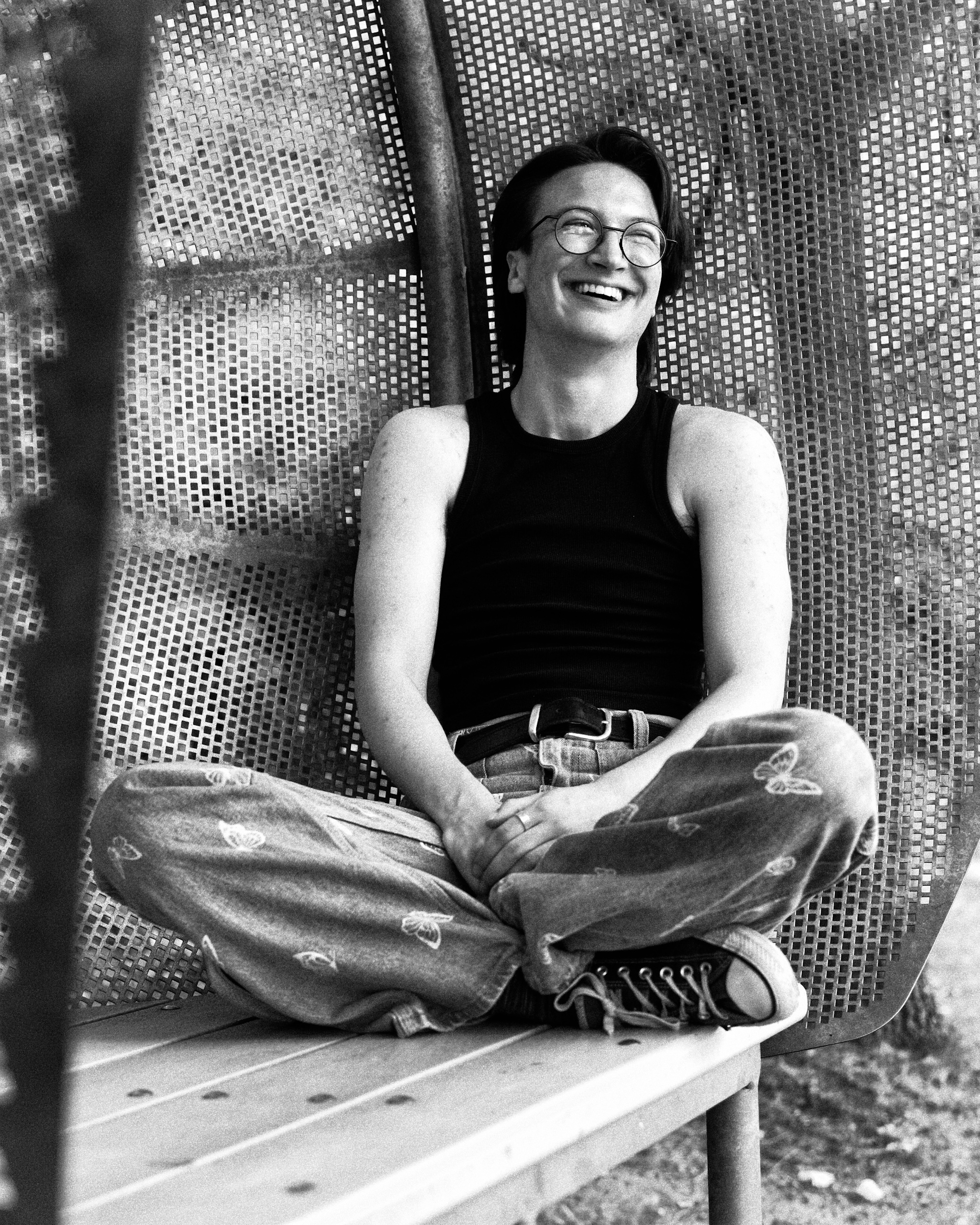 Monochrome portrait of a smiling adult male in eyeglasses and tank top, sitting cross-legged on a bench under a metal structure outdoors.