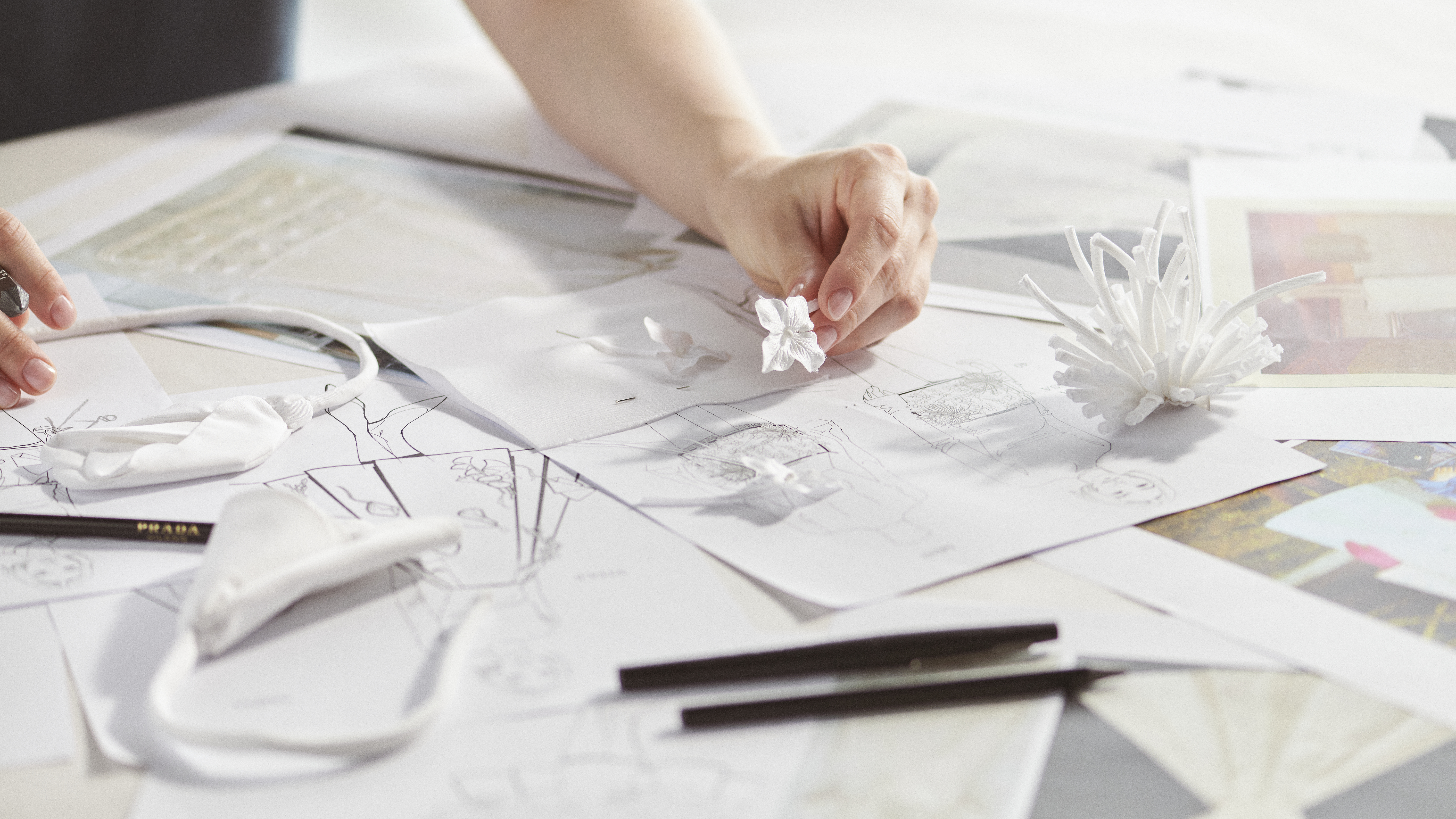 Close-up of a hand arranging small white floral accessories on top of fashion design sketches, with pencils and white fabric pieces.