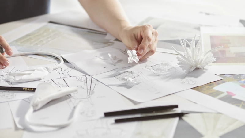 Close-up of a hand arranging small white floral accessories on top of fashion design sketches, with pencils and white fabric pieces.
