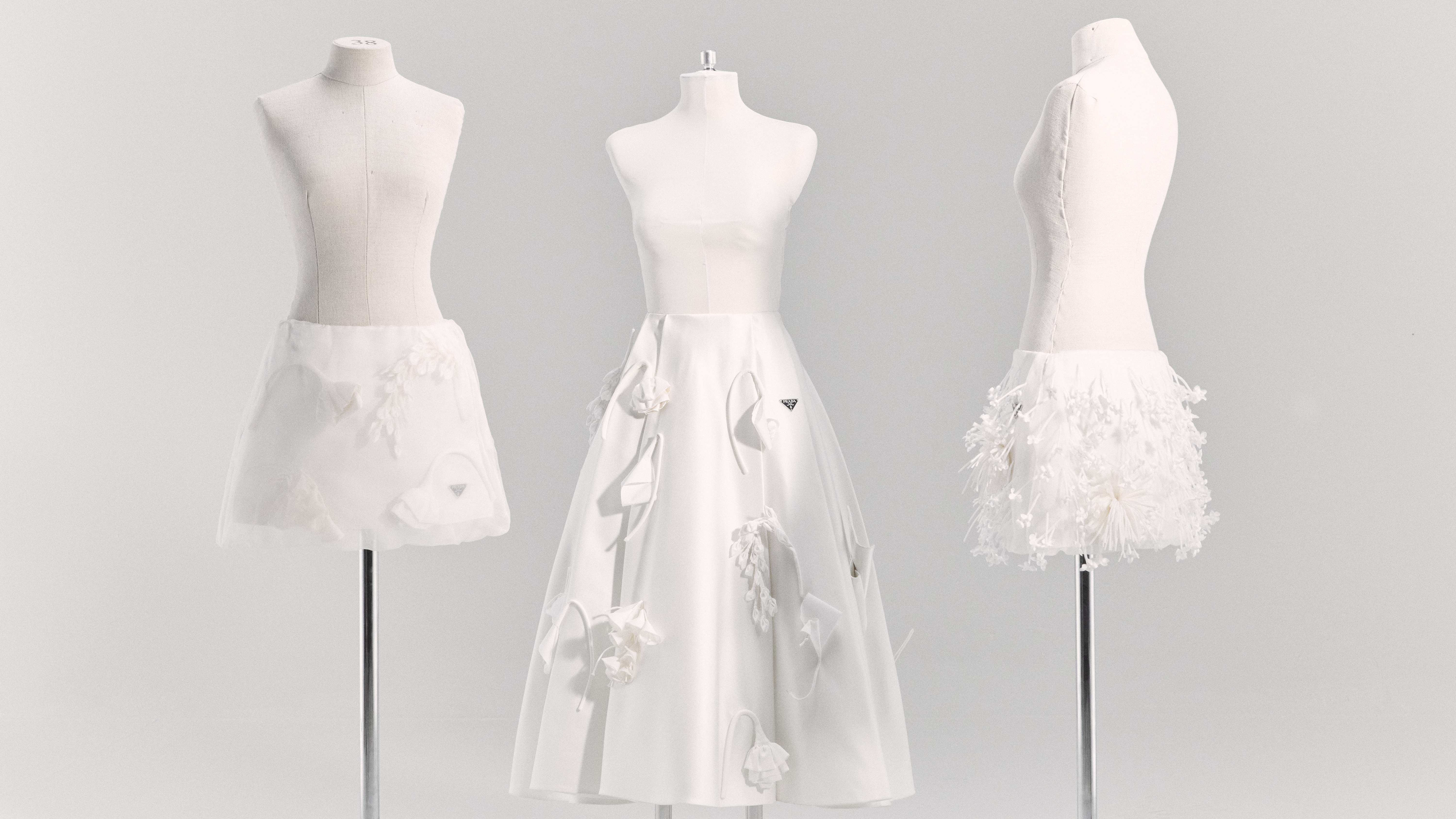 Three dress forms display white dresses with varying designs, set against a clean, bright backdrop in a fashion studio environment.