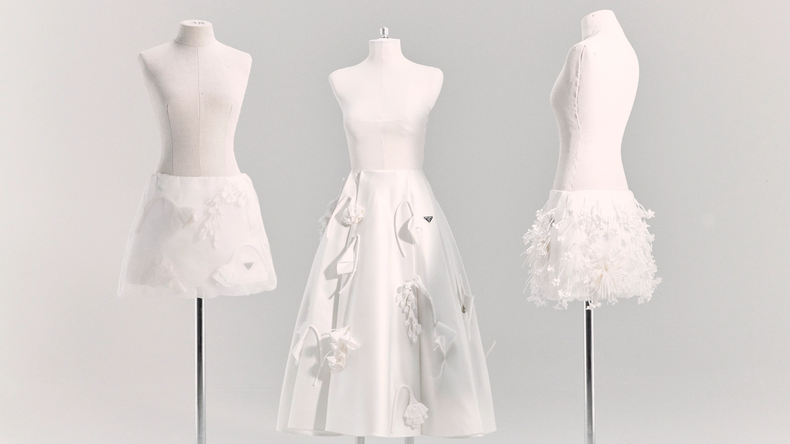Three white tailor mannequins displaying white Prada skirts with 3D floral embellishments against a pale seamless studio background.