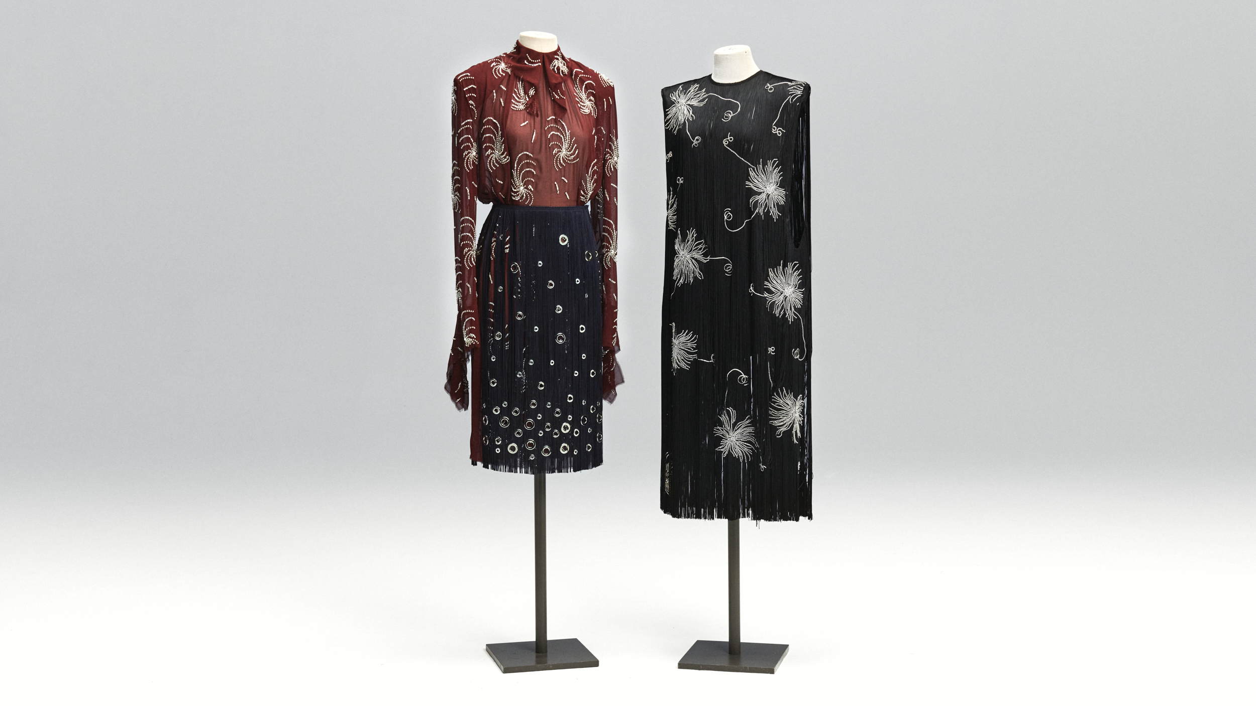 Two vintage-style dresses, one sheer red blouse with navy skirt and another black dress with silver floral patterns on display.