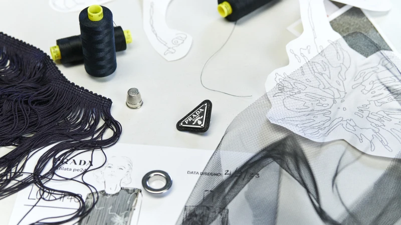 Fashion design elements, featuring thread spools, fabric swatches, Prada logo, thimble, fringe, and sketches on a white surface.