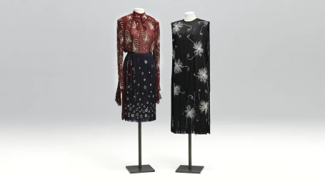 Two mannequins displaying an embroidered burgundy blouse with skirt and a black beaded fringe dress on a seamless studio background.