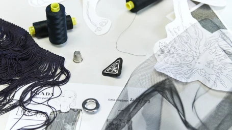 Fashion design atelier flat lay featuring a Prada triangle logo badge, black thread spools, fringe trim, sketches, and black mesh fabric.