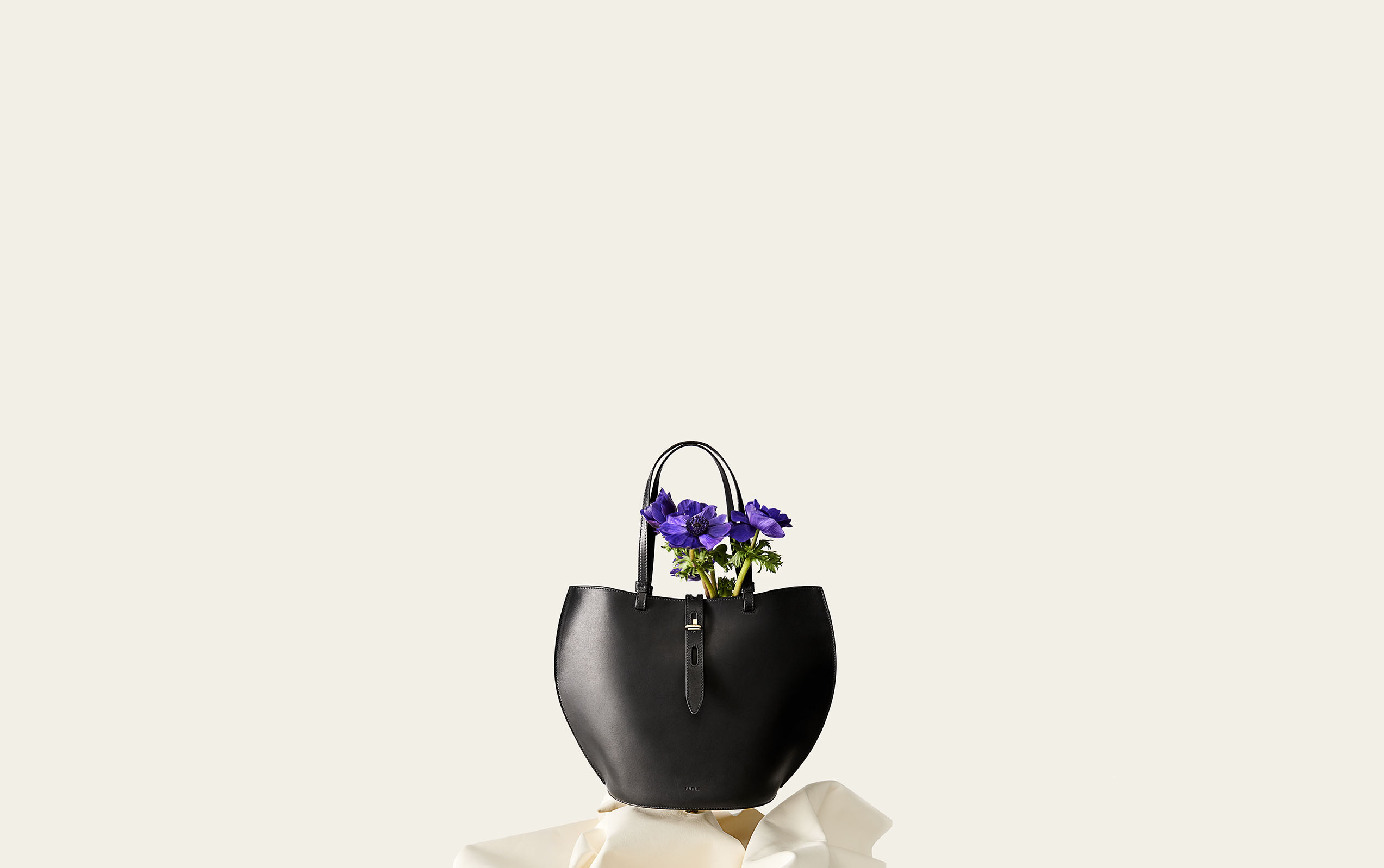 Black tote bag with purple anemone flowers inside, placed on white crumpled paper, set against a white seamless background.