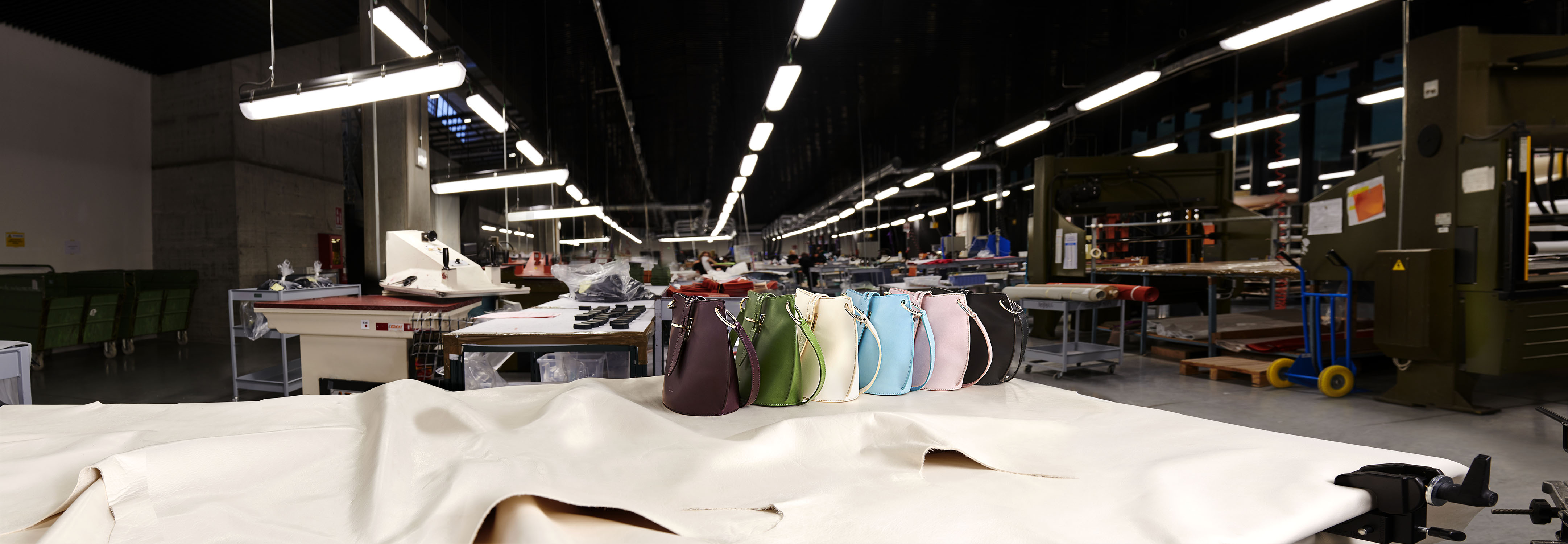Handbags in various colors lined up on a table in a workshop, showcasing a fashion collection in a manufacturing environment.
