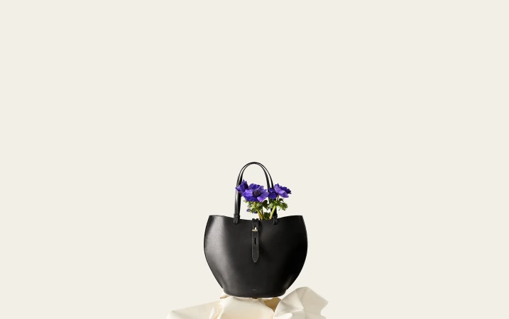 Black leather tote bag holding purple anemone flowers set against a minimal off-white background.