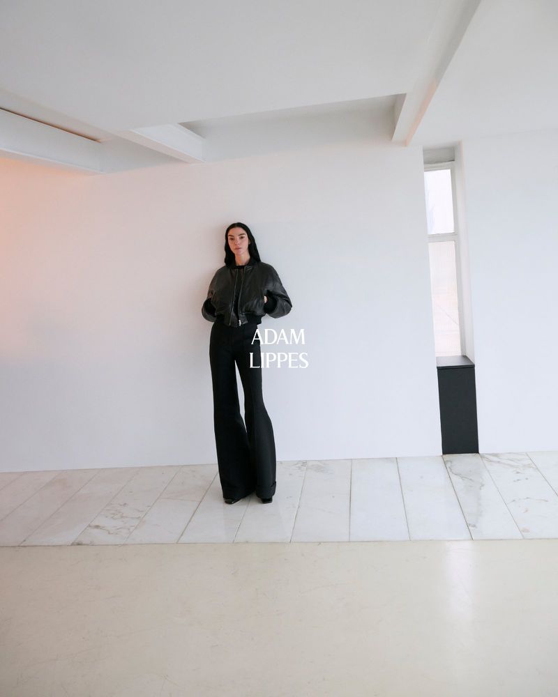A slender adult woman poses in a black bomber jacket and wide-leg pants against a white wall in a minimalist architectural space.