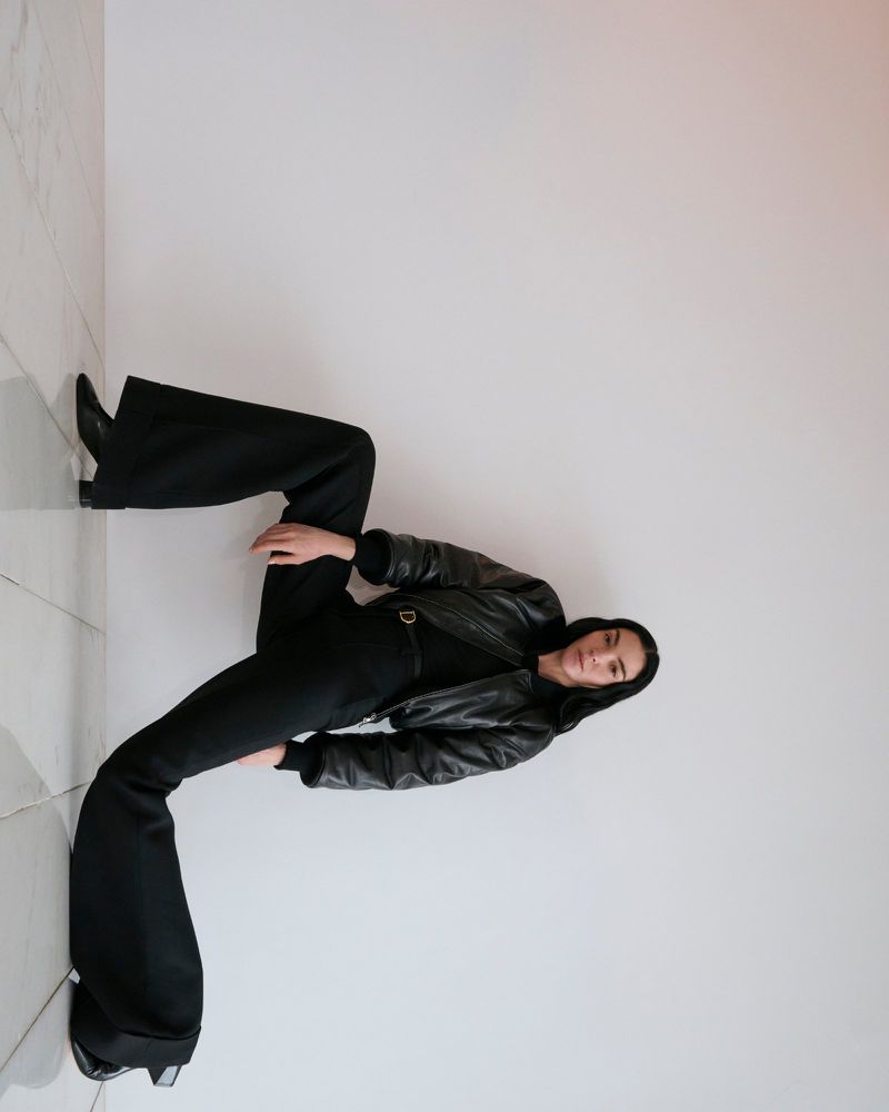 Woman in black outfit strikes a pose in a studio, wearing a leather jacket, wide leg pants, and heeled boots against a white wall.