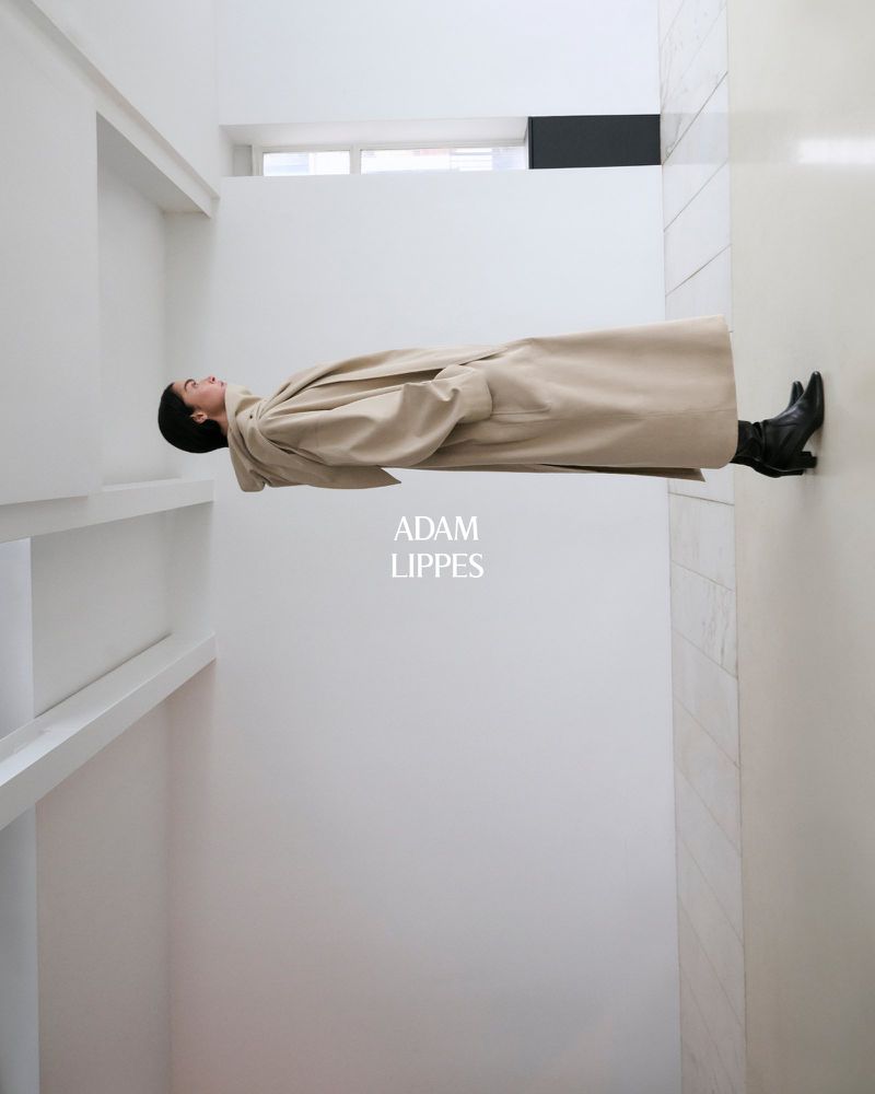 Fashion shot of a woman wearing a long trench coat and boots lying horizontally against the backdrop of a modern white interior.