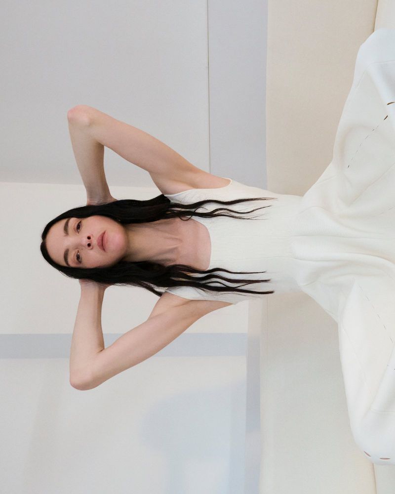An adult woman with long dark hair lies on her back in a white dress in a minimalist interior, posing with her hands behind her head.