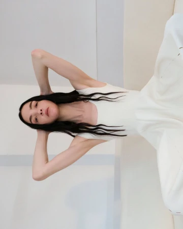 An adult woman with long dark hair lies on her back in a white dress in a minimalist interior, posing with her hands behind her head.