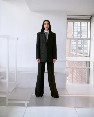 An adult female model stands in a light-filled interior wearing a black suit with wide-legged trousers and a striped tie. The styling is formal.