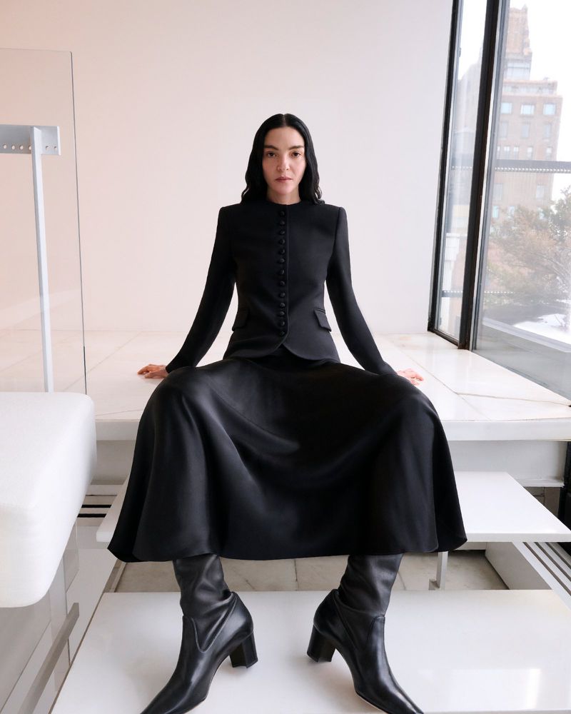 Elegant woman poses in black skirt suit and boots against a bright backdrop with a window and modern furniture, fashion shoot.