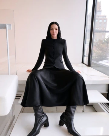 Elegant woman poses in black skirt suit and boots against a bright backdrop with a window and modern furniture, fashion shoot.