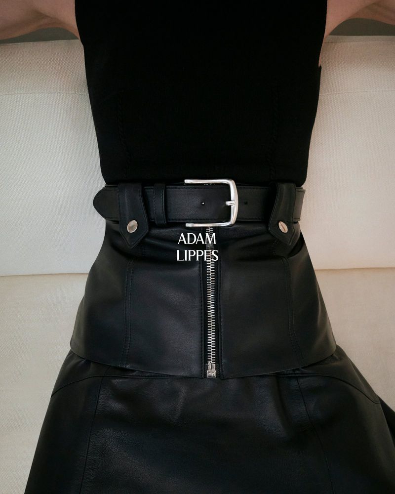 Close-up of a woman wearing a black leather skirt and matching belt with a silver buckle, against a neutral background.