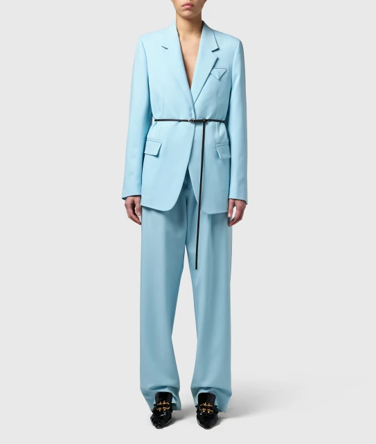 Woman in light blue pantsuit with black belt standing in front of a plain, light gray studio backdrop for a fashion editorial shoot.