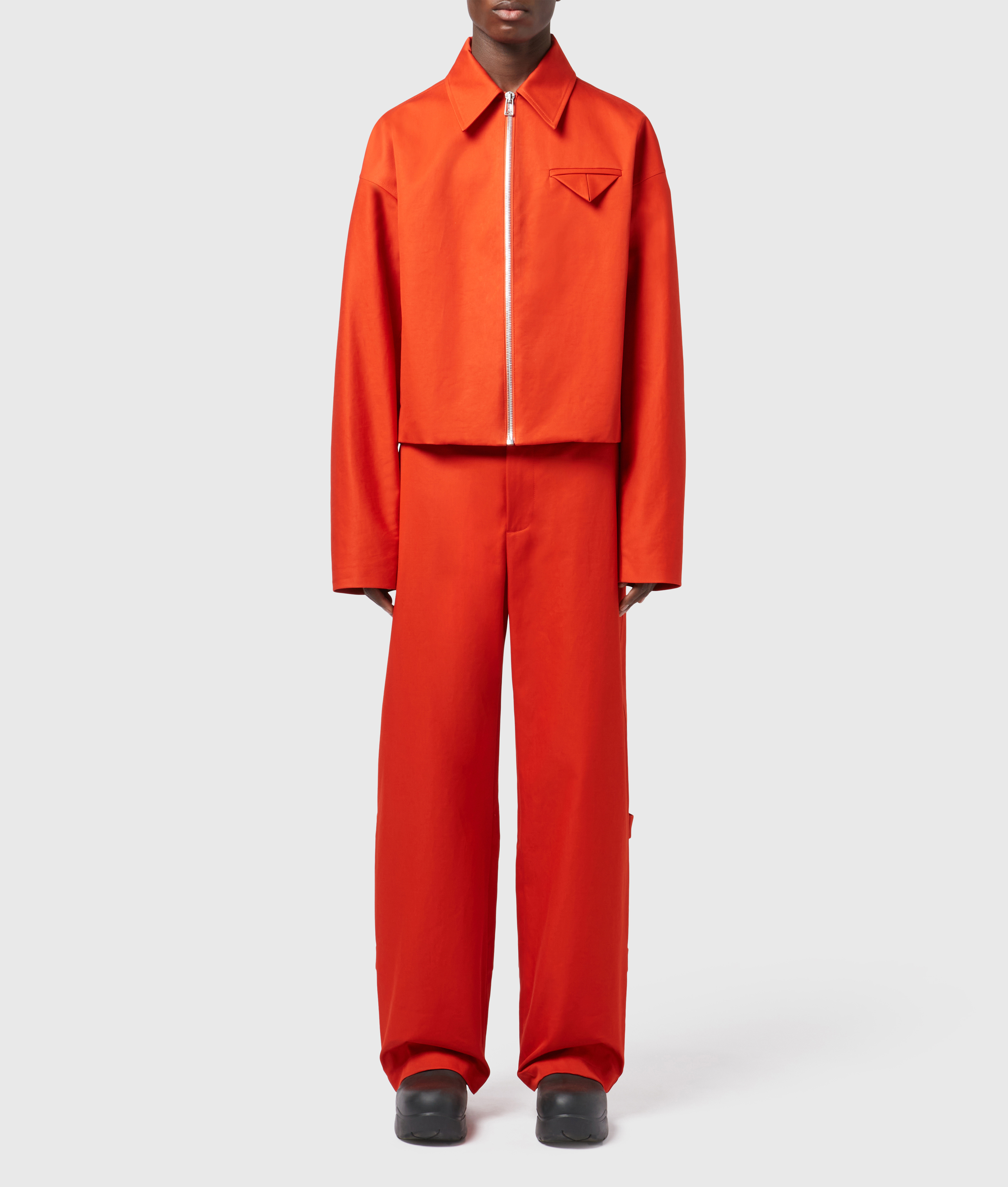 Person wearing a bright orange jacket with zipper and matching orange wide leg pants. Fashion shot in a neutral studio setting.