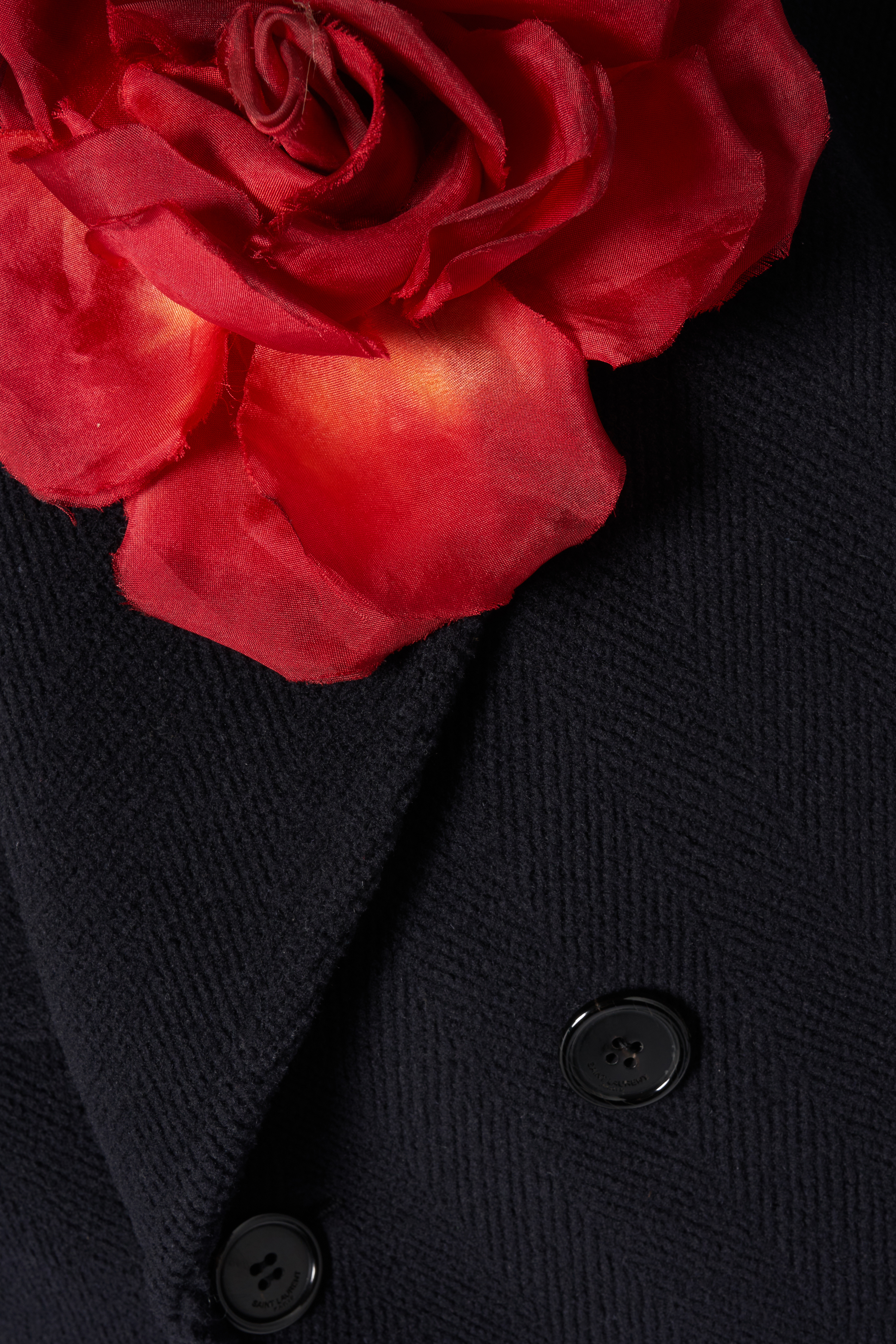 Close-up of a large red rose pinned to a navy blue wool herringbone coat, secured with black buttons for a formal or elegant look.