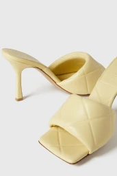 A pair of pale butter yellow quilted leather high-heeled mules with square open toes displayed on a white seamless background.