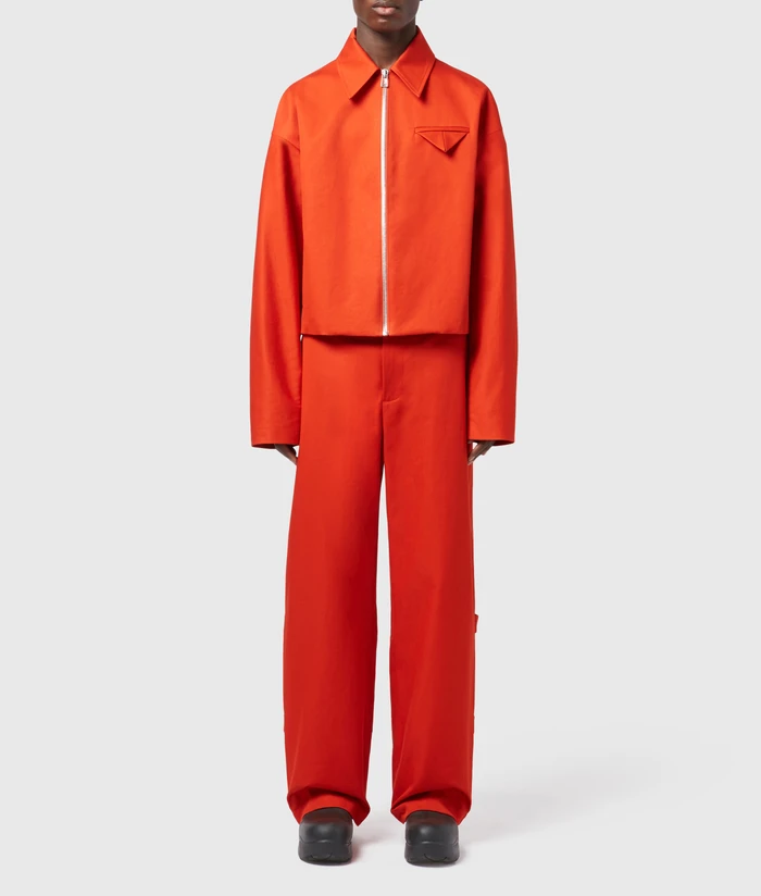 Model wearing a bright red-orange cropped zip-up jacket and matching wide-leg trousers with chunky black shoes against a grey background