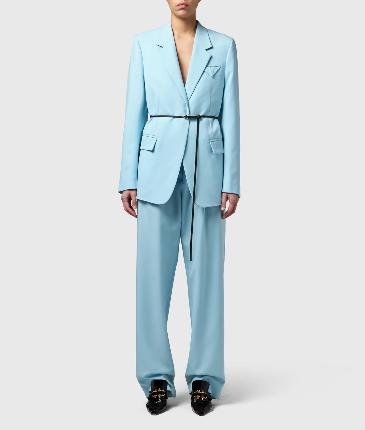 Model wearing a light blue tailored pantsuit with a thin black belt and black shoes against a white background.