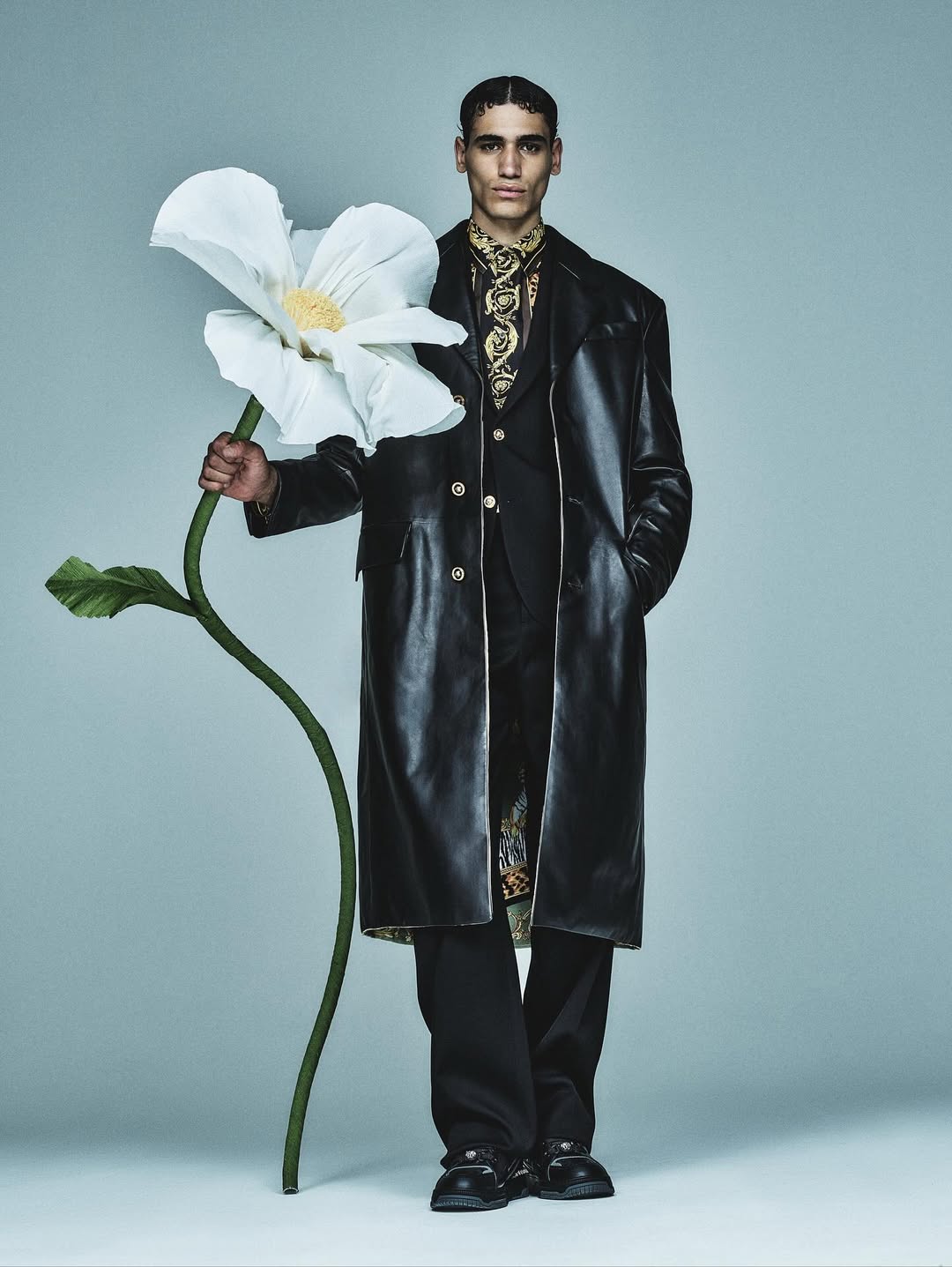 Adult male model stands in a black leather coat, holding a large, white flower, in a studio fashion shot with a light grey background.