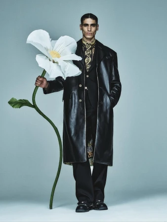 Adult male model stands in a black leather coat, holding a large, white flower, in a studio fashion shot with a light grey background.