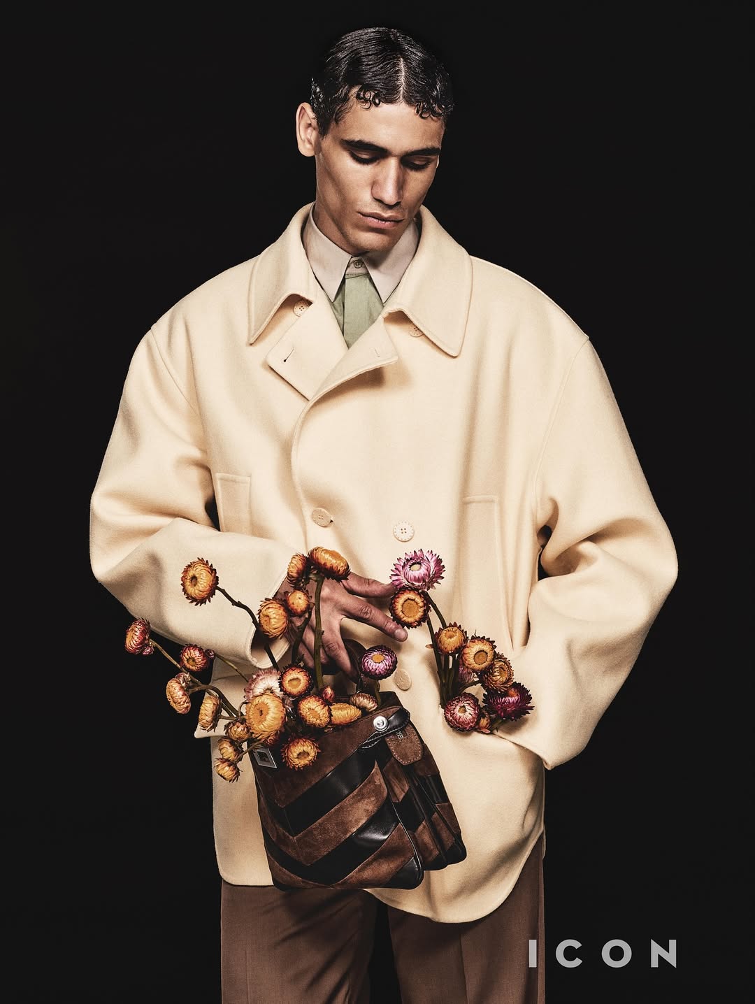 Editorial fashion shot of a young adult man wearing a beige overcoat and holding a purse with dried flowers against black backdrop.
