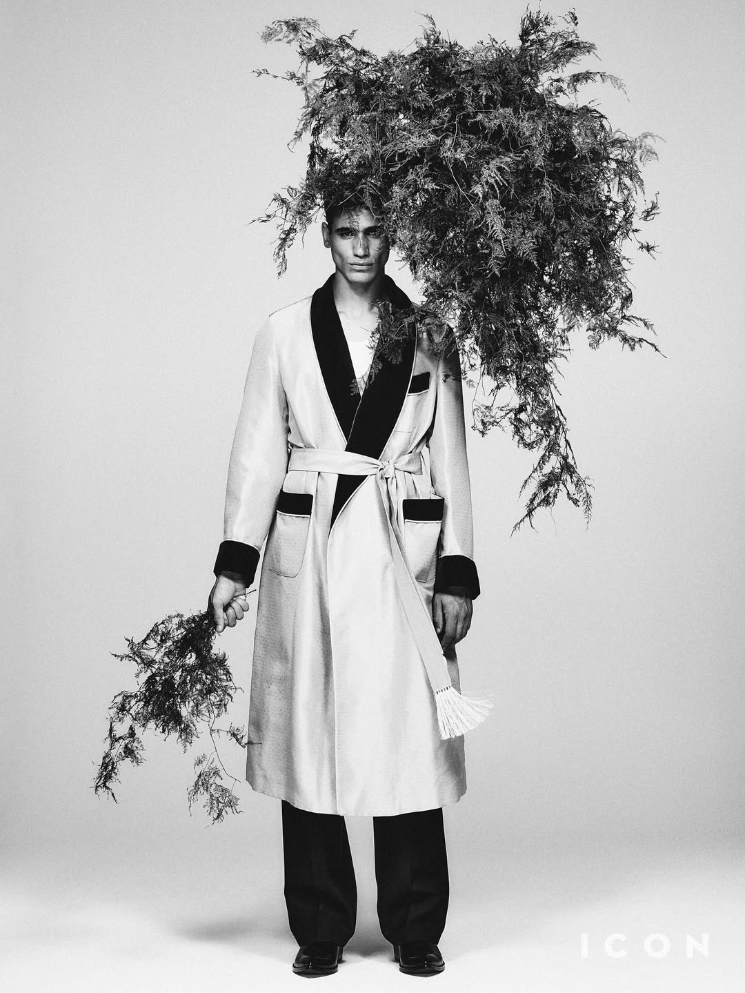 Black-and-white portrait of a man in a robe, adorned with greenery on his head and hand, against a light background. Fashion editorial.